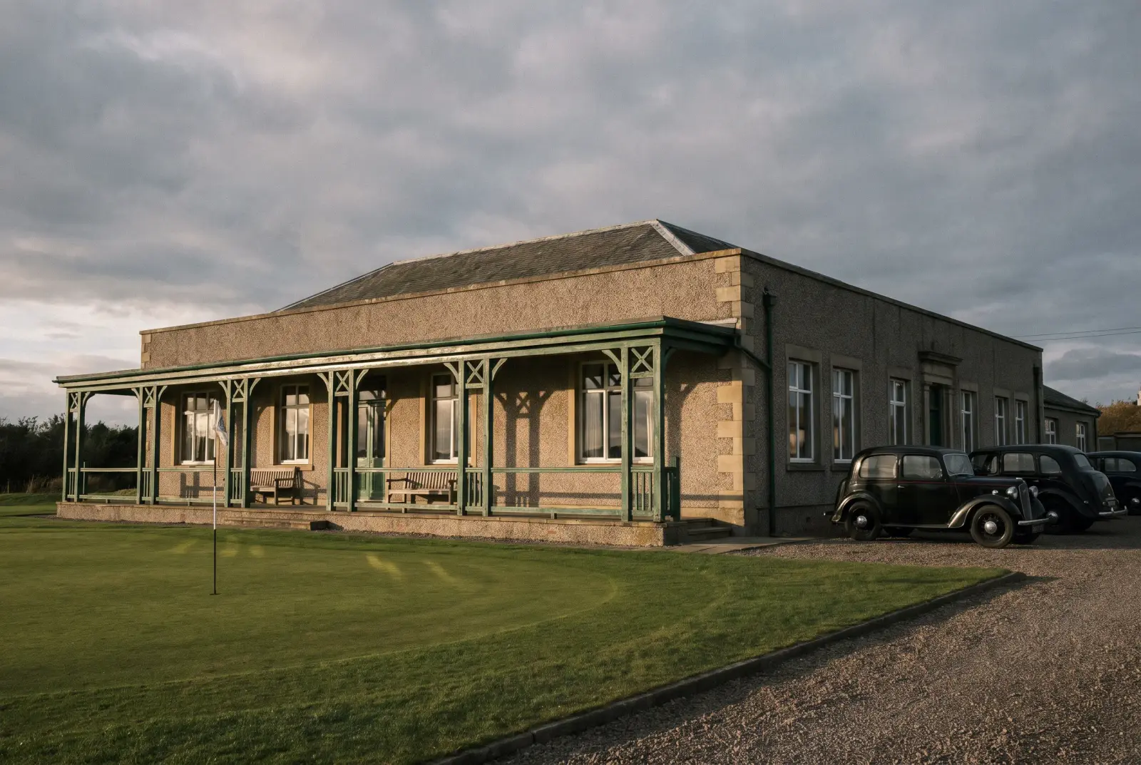 The clubhouse from the eighteenth green: a low stone building with a green-painted veranda, vintage cars parked outside