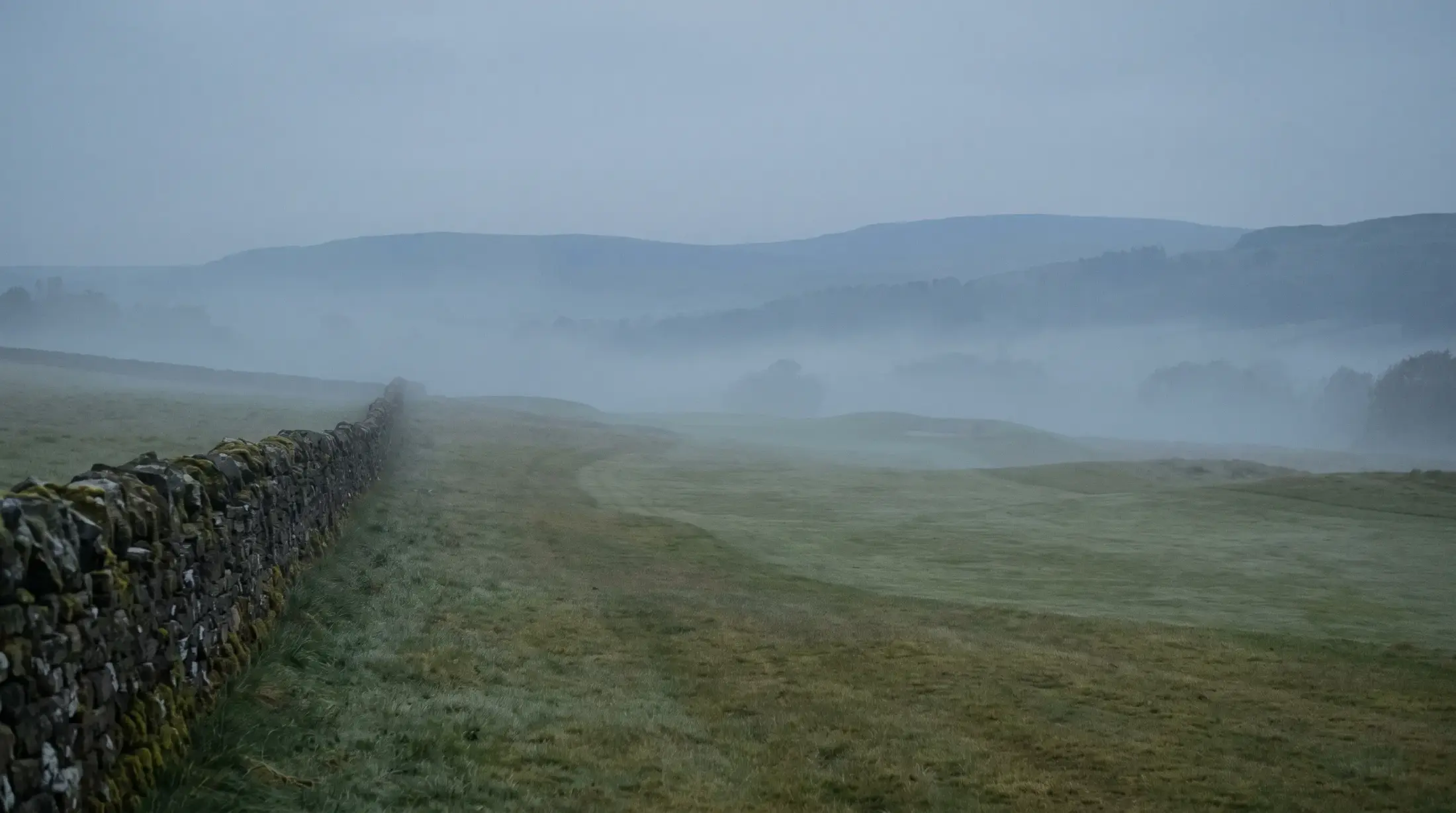 The valley before sunrise, mist in the folds of the ground, a dry stone wall running toward the dark hills