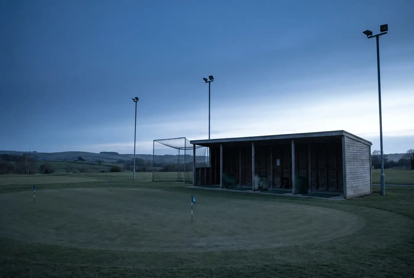 The practice ground at dusk, floodlit bays and a netted driving area beside three practice greens